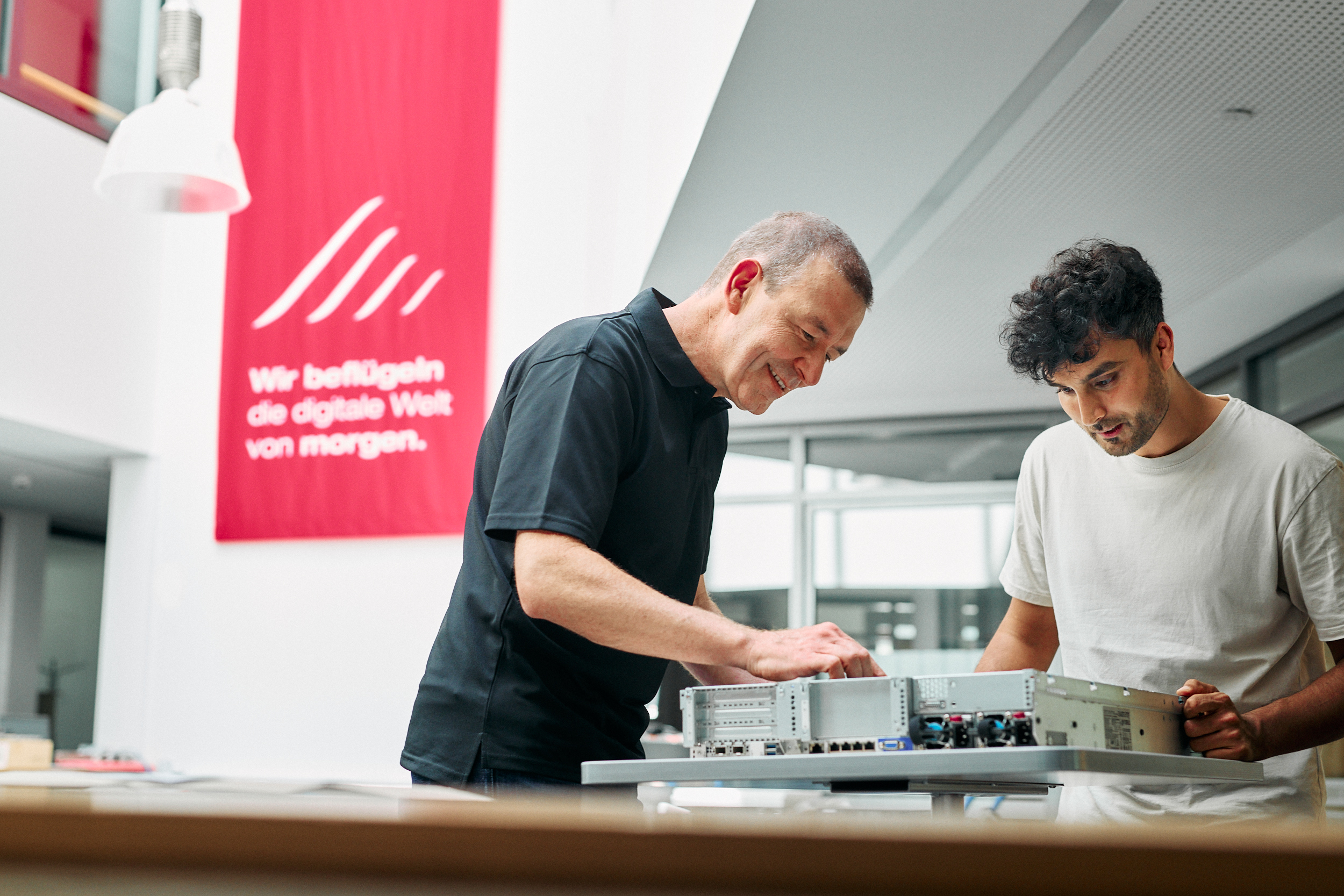 Zwei Mitarbeitende schauen gemeinsam in eine geöffnete Hardware, im Hintergrund sieht man einen roten banner mit der IBYKUS Vision